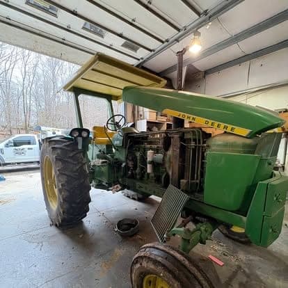 A green John Deere tractor pulled into the shop with the side panels and hood removed for engine work.