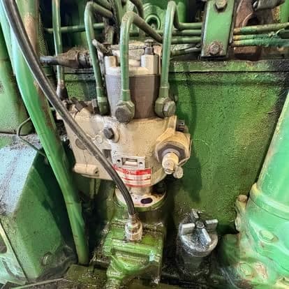 The hydraulic pump assembly and steel lines mounted on the side of a green tractor engine.