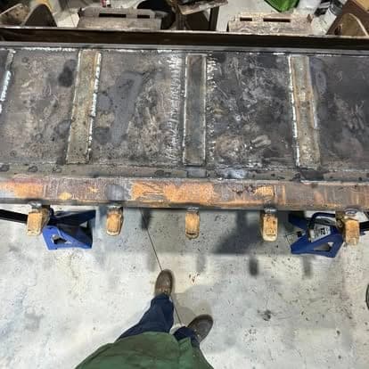 Fresh weld beads running along wear plates laid onto a steel cutting edge, clamped down on the shop bench.